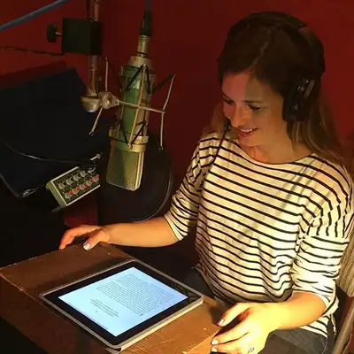 A person reads a script from an iPad while seated in a recording studio, wearing headphones and positioned near a microphone.