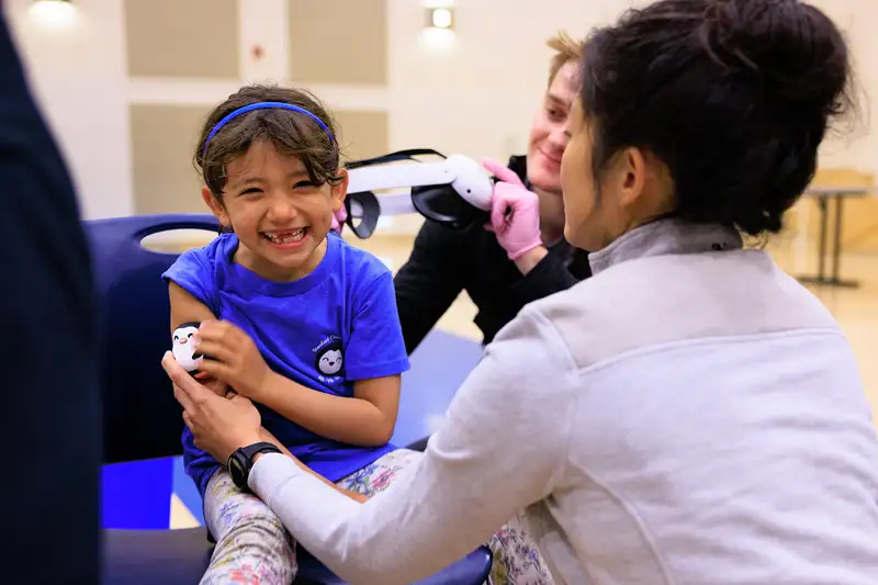 A health care worker in gloves is preparing to give a vaccination to a young child sitting in a chair while another adult assists.