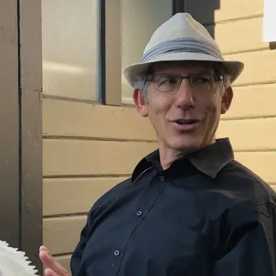A man in a dark blue shirt and white hat sits near a door, gesturing with his hand as he engages in conversation.