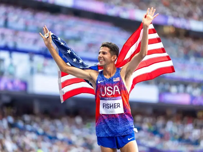 Grant Fisher raises both arms in celebration with an American flag draped over his shoulders, smiling in a packed stadium at the Paris 2024 Olympic Games.