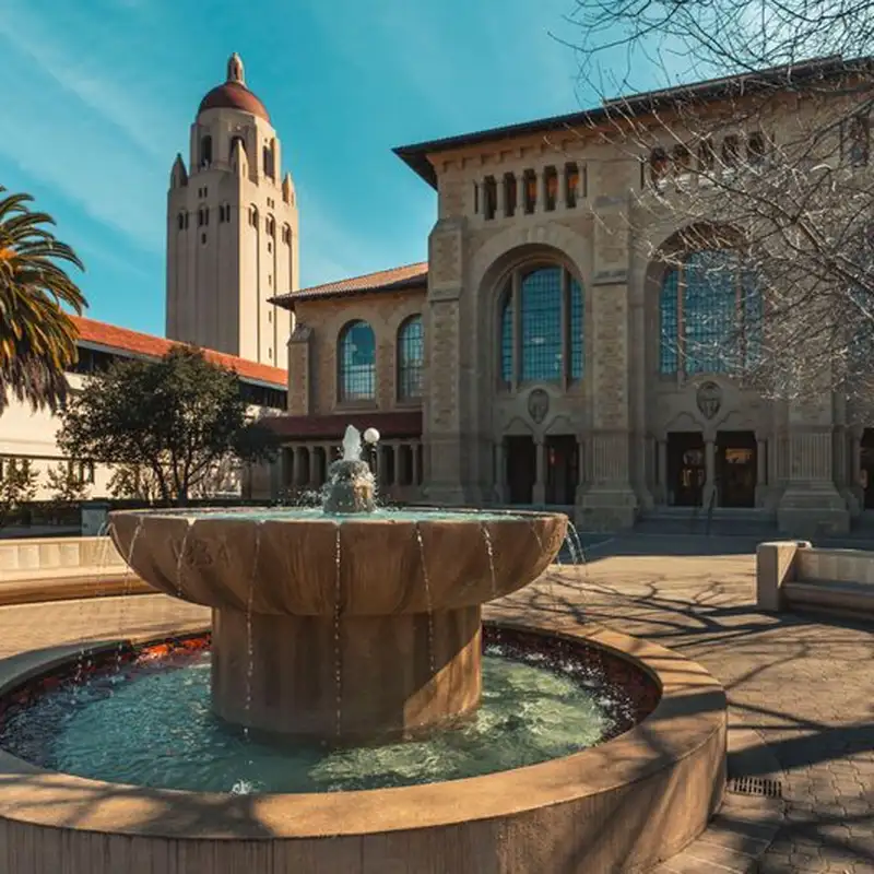 A stone fountain in the foreground frames a view of Stanford's sandstone buildings and Hoover Tower rising against a bright blue sky.