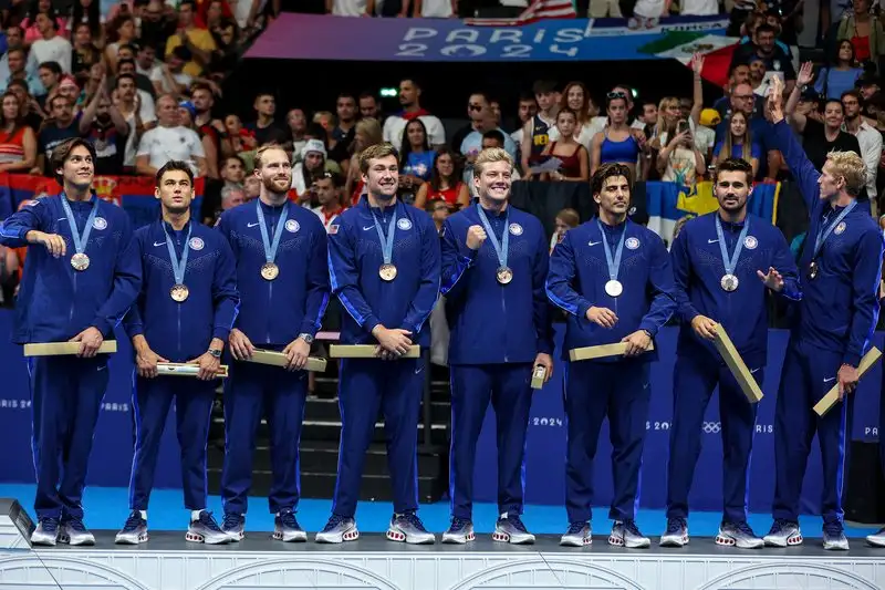 The US men's water polo team stands in a row in blue Team USA tracksuits wearing their bronze medals on a podium at the Paris 2024 Olympic Games.