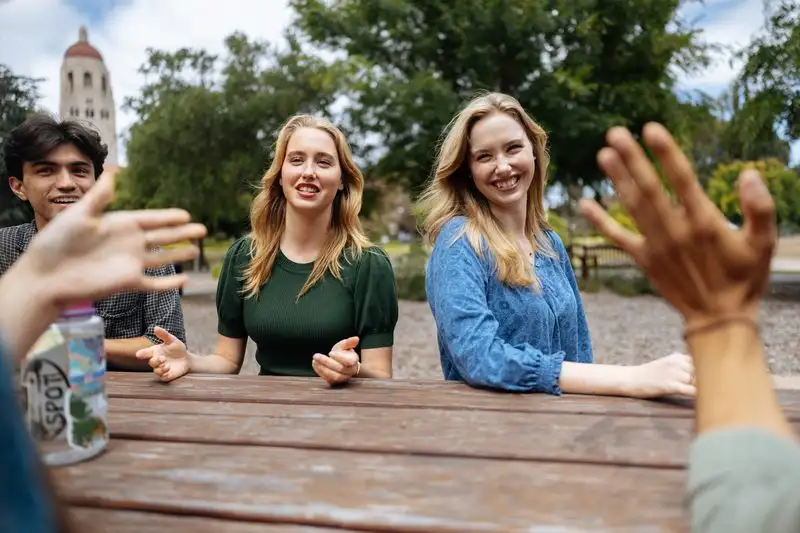 Katelyn and Lorelei Santa Maria having a lively conversation with their friends at a picnic table outdoors, with trees and the Hoover Tower visible in the background.