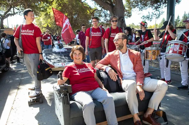 Provost Drell and Andrew Luck ride on a motorized couch through campus, solar car behind them, band to the side