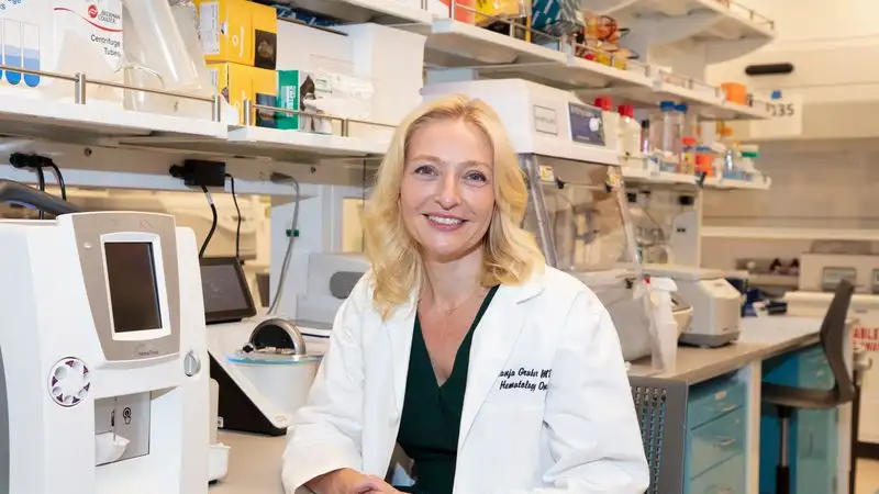 Dr. Tanja Gruber, a hematology-oncology physician in a white coat, smiles while seated at a lab bench surrounded by scientific equipment and supplies.