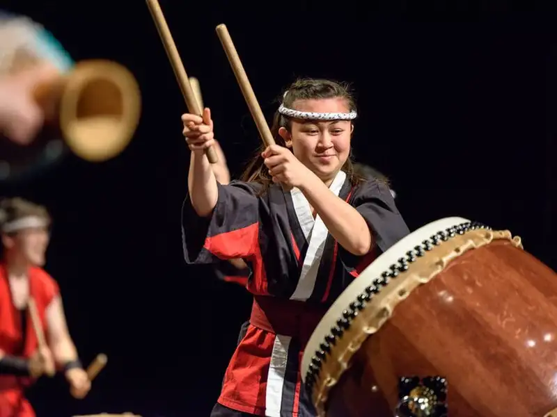 A taiko drummer in a black and red happi coat and headband raises both drumsticks mid-strike at a large drum during a performance on a dark stage.