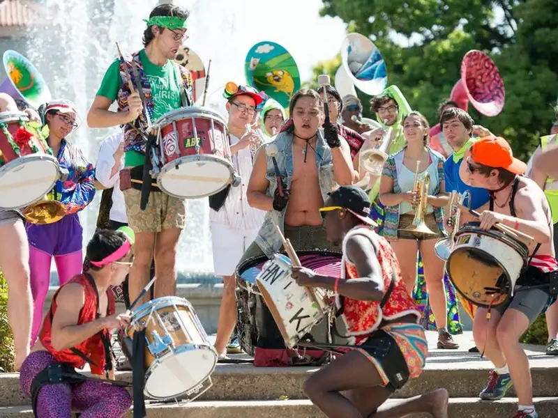 Members of a marching band in colorful, eclectic outfits energetically play drums, trumpets, and sousaphones near a fountain during an outdoor admit-weekend performance.