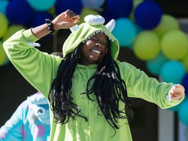A person in a green one-eyed monster onesie dances joyfully with arms raised under an arch of green and blue balloons at a student move-in event.