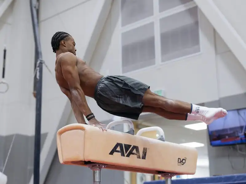Gymnast Khoi Young performs a pommel horse routine, body extended horizontally and legs straight out, supporting himself on one arm in a training gym.