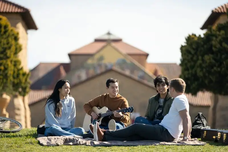 Four students sit on a blanket on a sunny lawn, one playing acoustic guitar, with a Stanford sandstone building behind them.