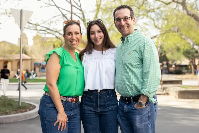 Three people, two parents and a young woman, smile together outdoors on a tree-lined campus walkway during a university family day.