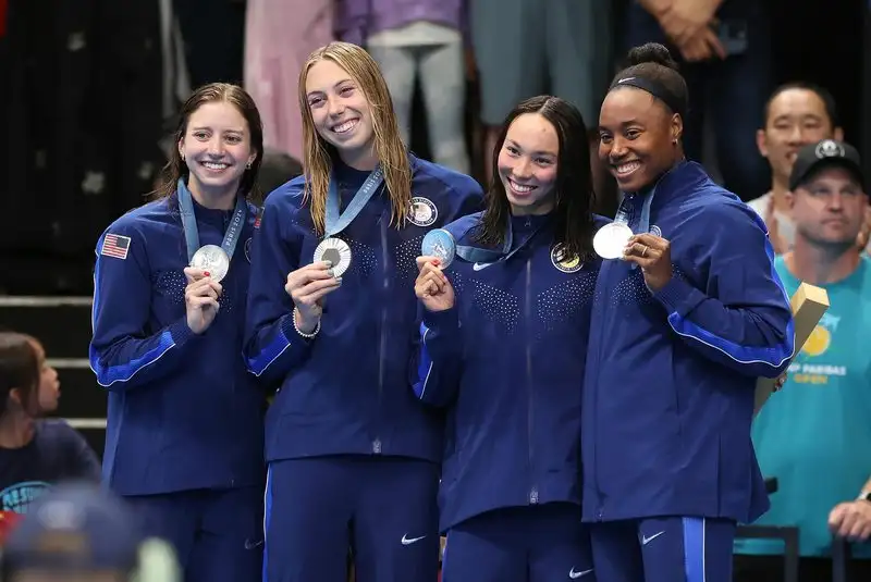 Four smiling US Olympic Team swimmers in blue jackets hold up their silver medals together at the Paris 2024 Olympic Games.
