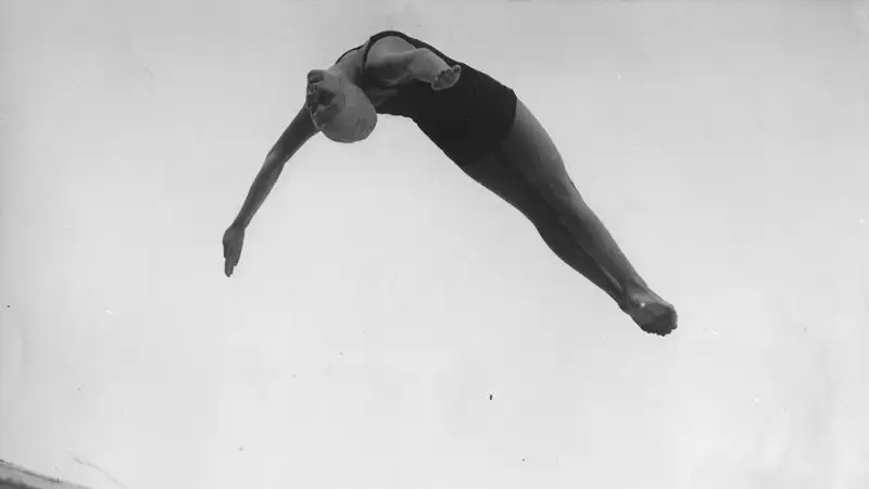 Marjorie Gestring arches through the air mid-dive above a diving board against a pale sky, in this black-and-white historical photo.