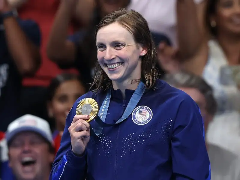 Katie Ledecky smiles and holds up her gold medal, wearing a blue Team USA jacket, in front of a cheering crowd at the Paris 2024 Olympic Games.