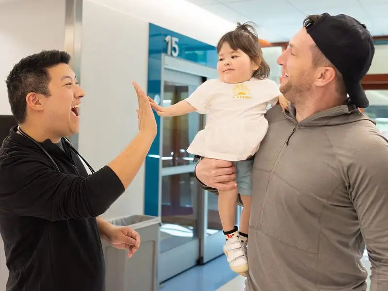 A dad holds his toddler girl, who is giving a high five to a man wearing a black shirt