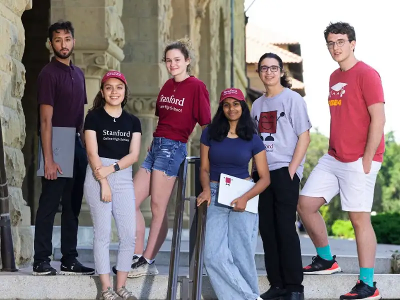 A group of six students poses on steps outside a building, wearing casual clothing and Stanford-themed apparel, smiling for the camera.