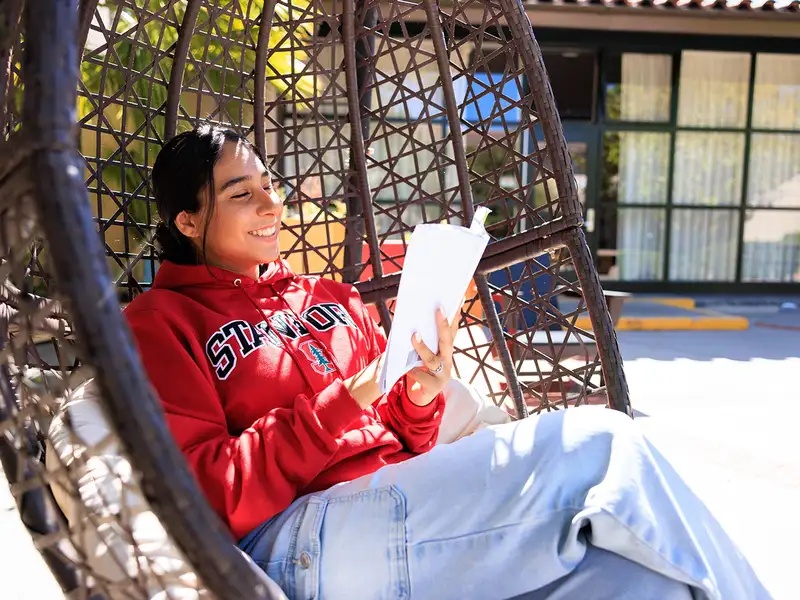 A student in a red hoodie relaxes in a hanging chair, reading from a notebook while enjoying a sunny outdoor setting.