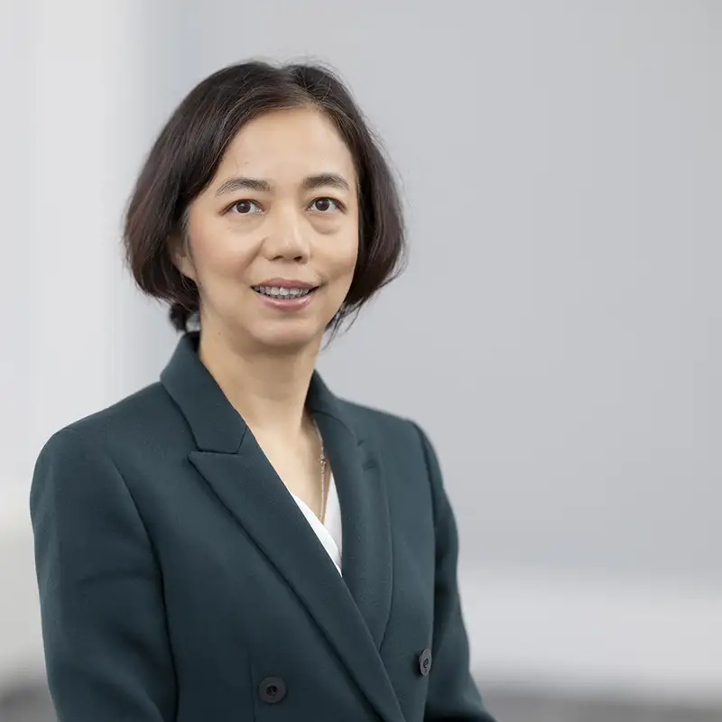 Fei-Fei Li wearing a dark green blazer over a white top, smiling against a blurred indoor background.