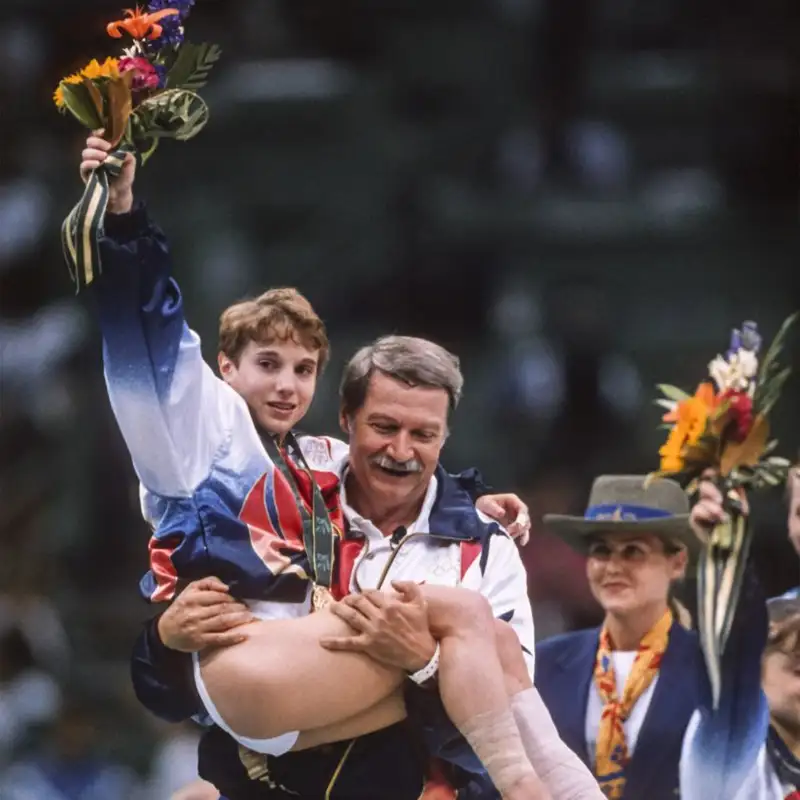 Gymnast Kerri Strug, holding a bouquet aloft and wearing her gold medal, is carried across the arena floor by her coach at an Olympic medal ceremony.