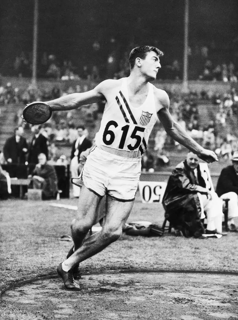 Bob Mathias, wearing a USA jersey numbered 615, winds up to throw the discus during a track and field competition, in this black-and-white historical photo.