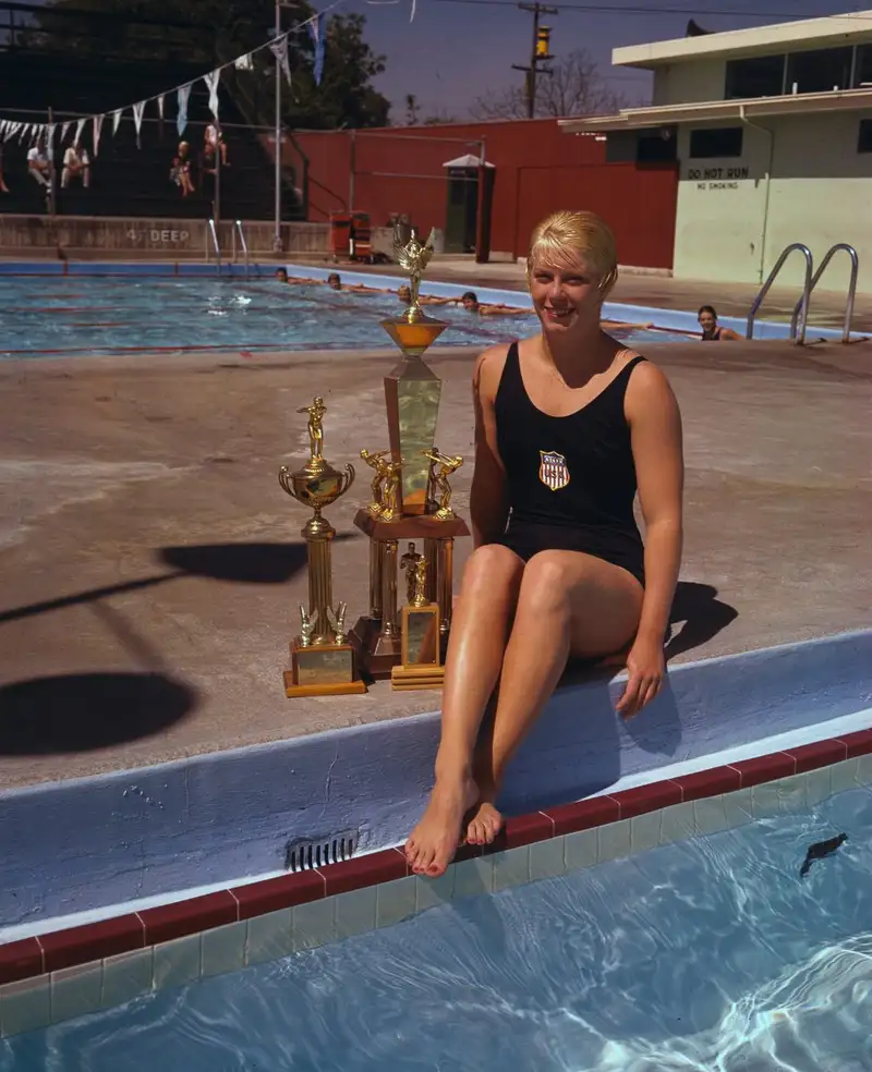 Swimmer Chris von Saltza, in a dark USA team swimsuit, sits smiling at the edge of an outdoor pool beside three large trophies on a sunny day.