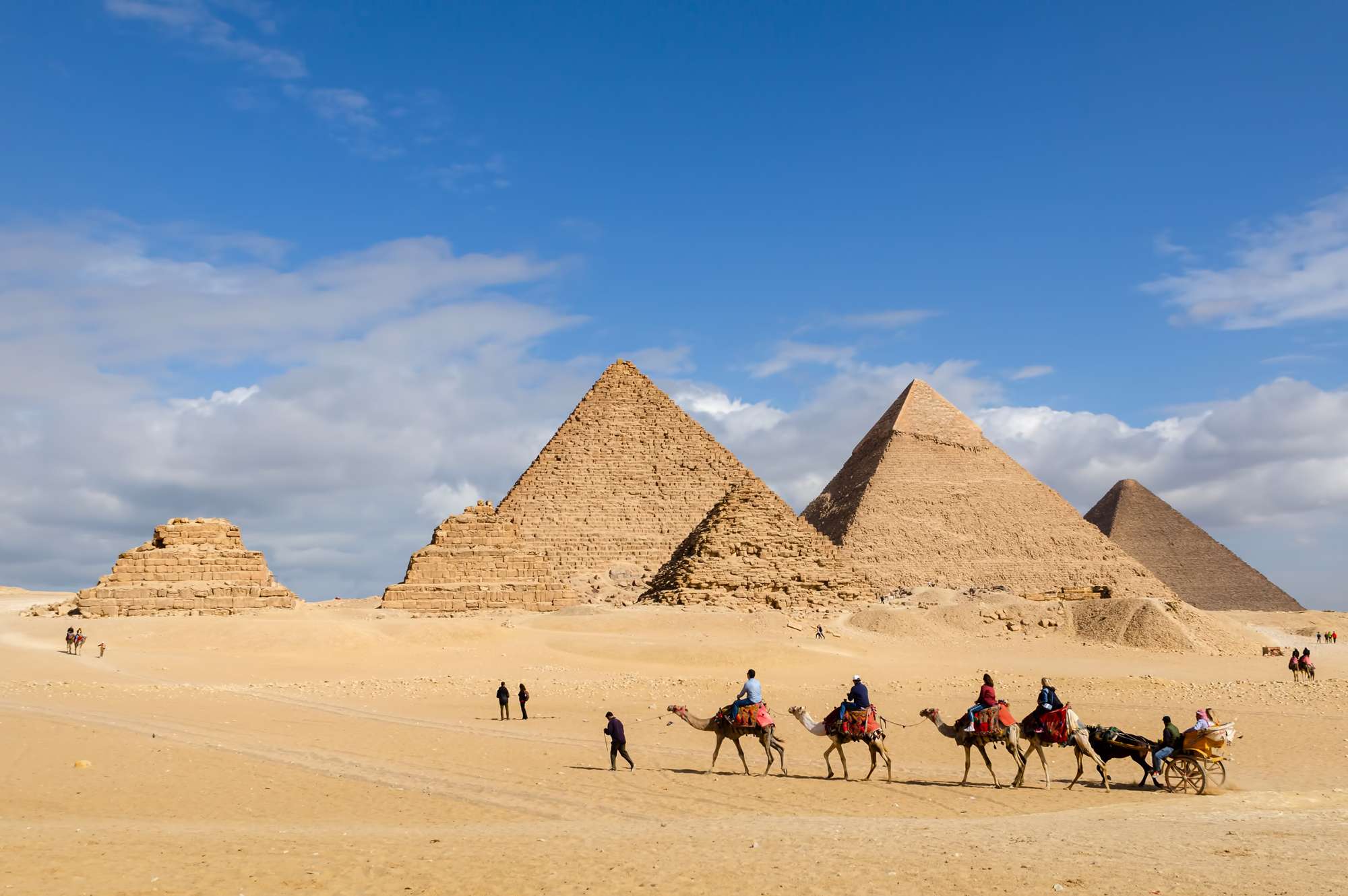 caravan of visitors riding camels with pyramids in distance