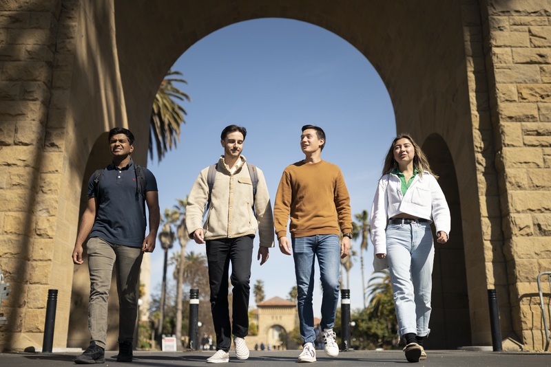 Four students walk in a line under a Stanford arch