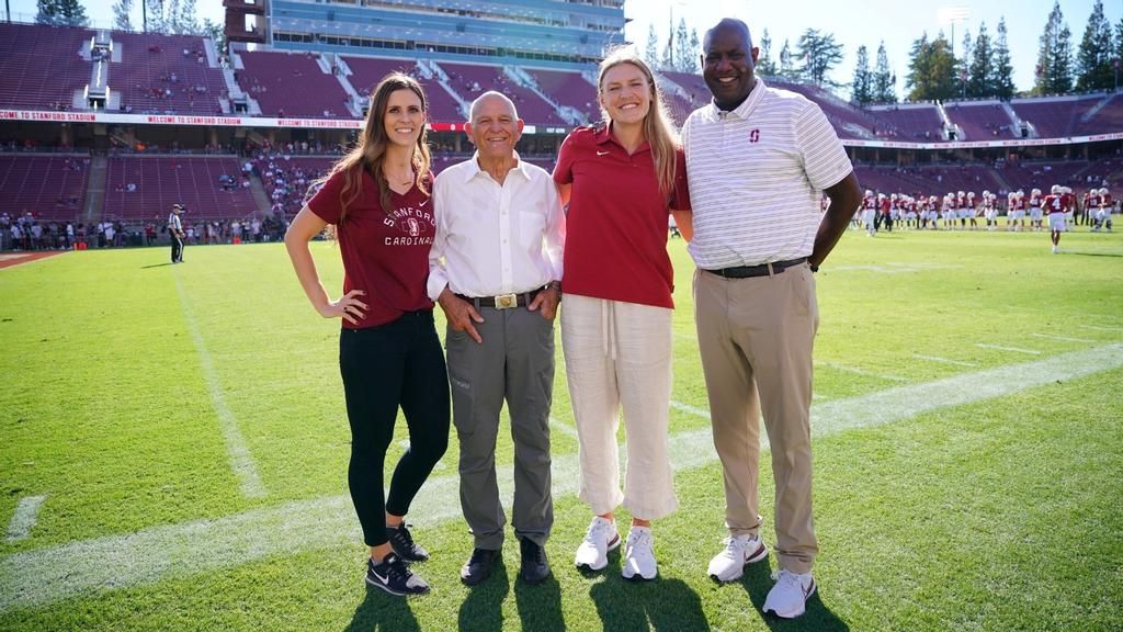 Four people standing together and smiling on the sidelines at Stanford Stadium
